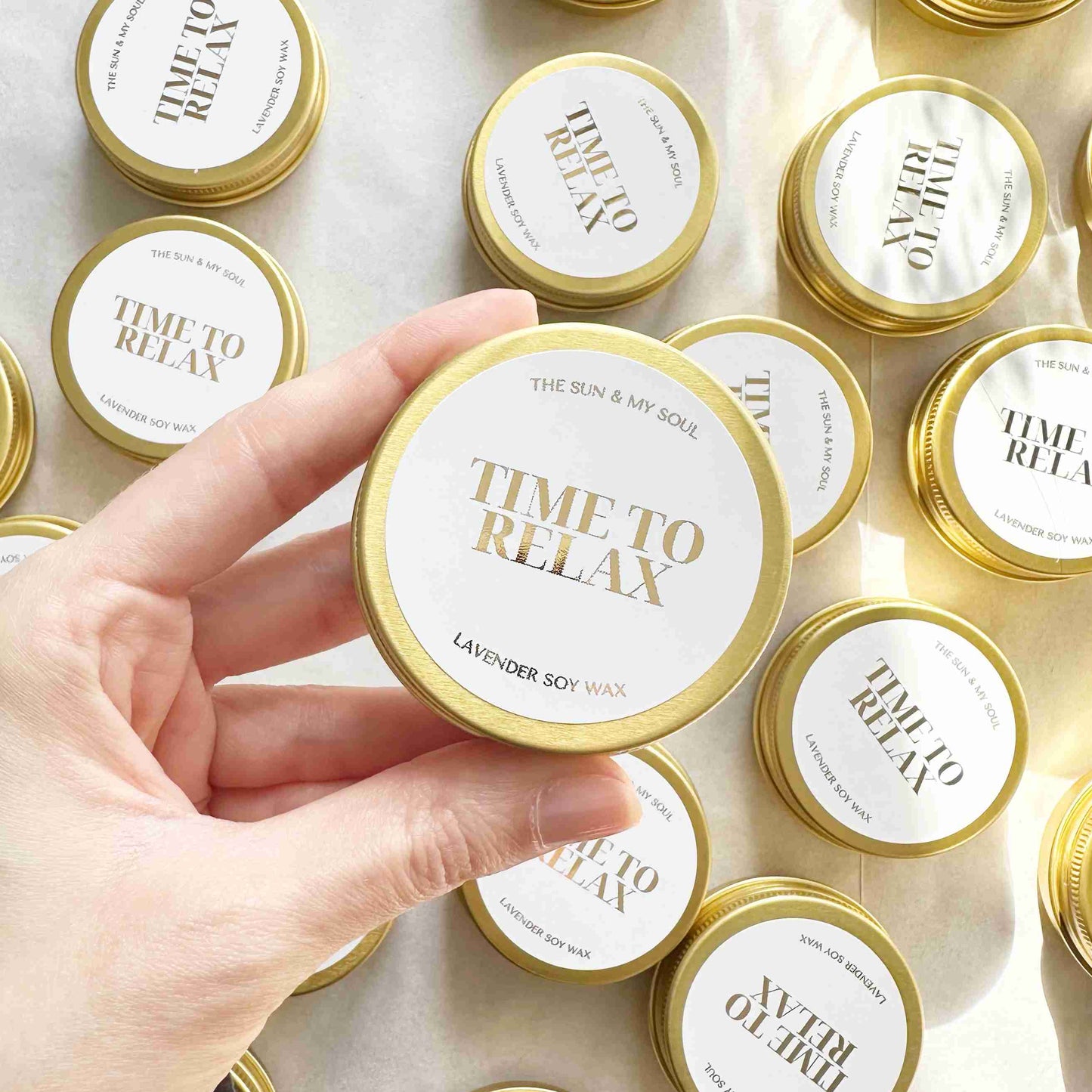 Hand holding a 'Time to Relax' candle with more candles in the background on a light surface.