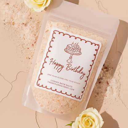 Packaged bath salts with a 'Happy Birthday' label, surrounded by white roses on a beige background