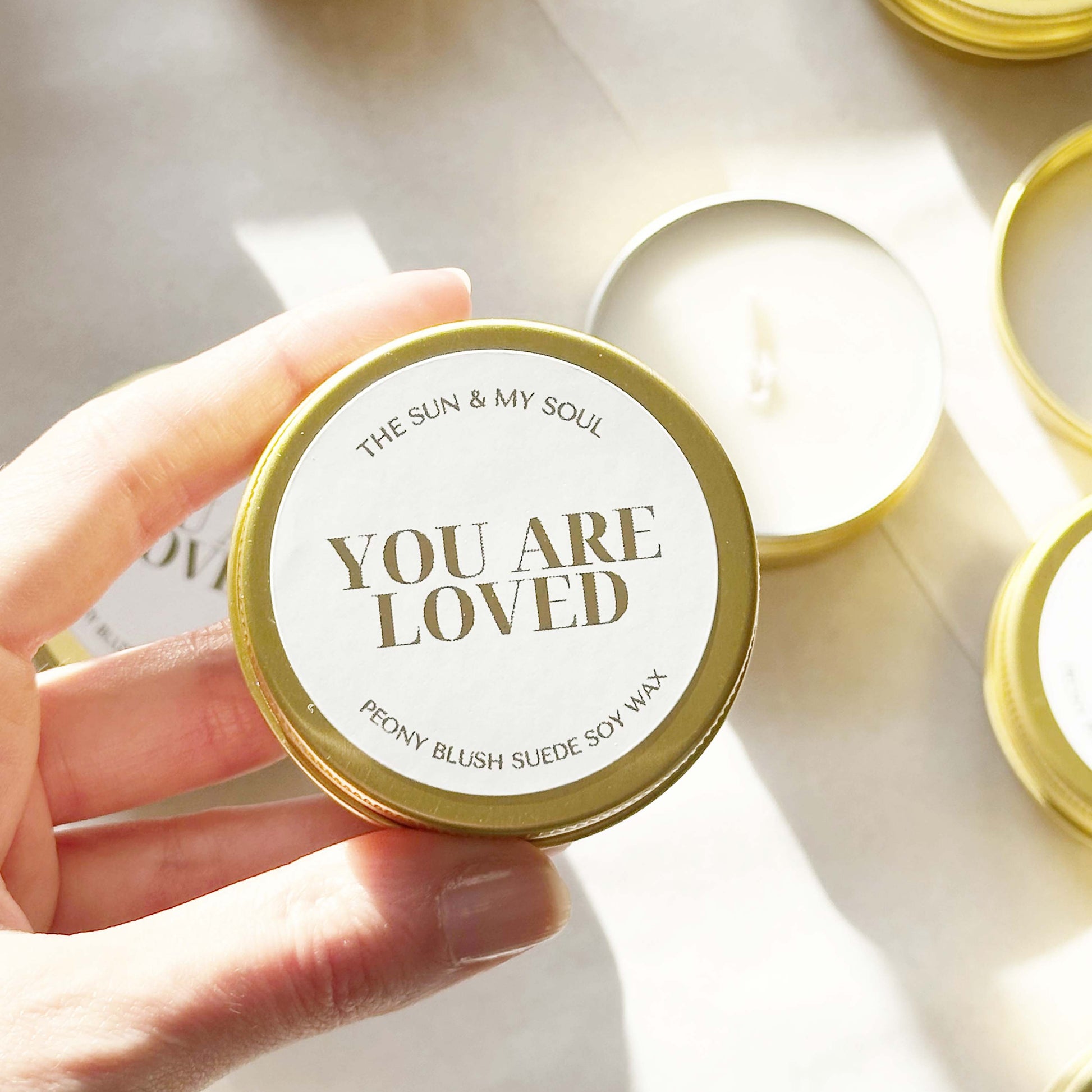 Candle with 'You Are Loved' lid held by a hand, surrounded by more candles on a light background