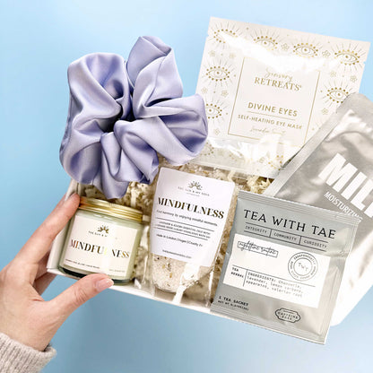 Hand holding a set of self-care products including a scrunchie, candle, and tea bags on a light blue background.