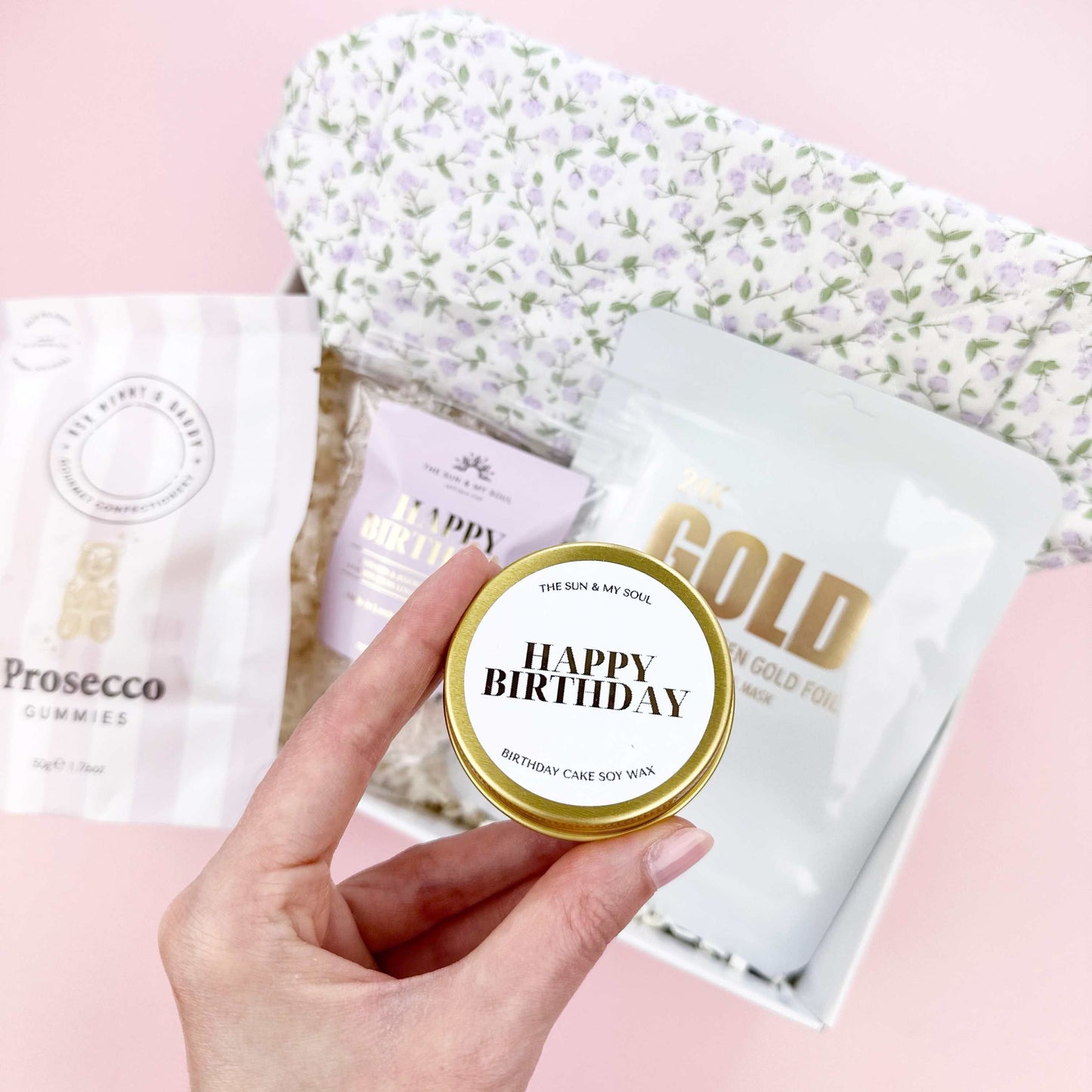 Hand holding a 'Happy Birthday' cake slice with more packages on a pink surface