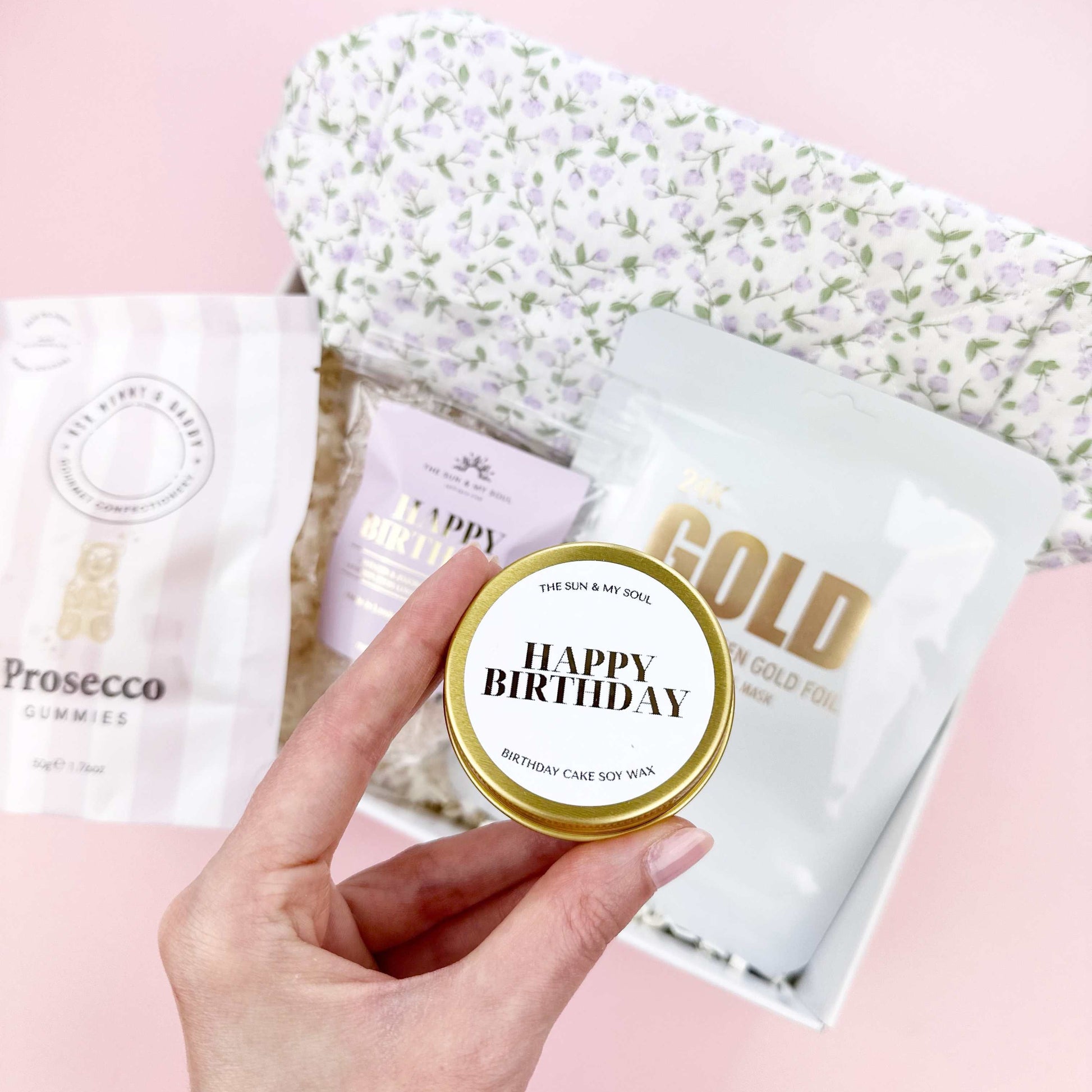 Hand holding a 'Happy Birthday' cake slice with more packages on a pink surface