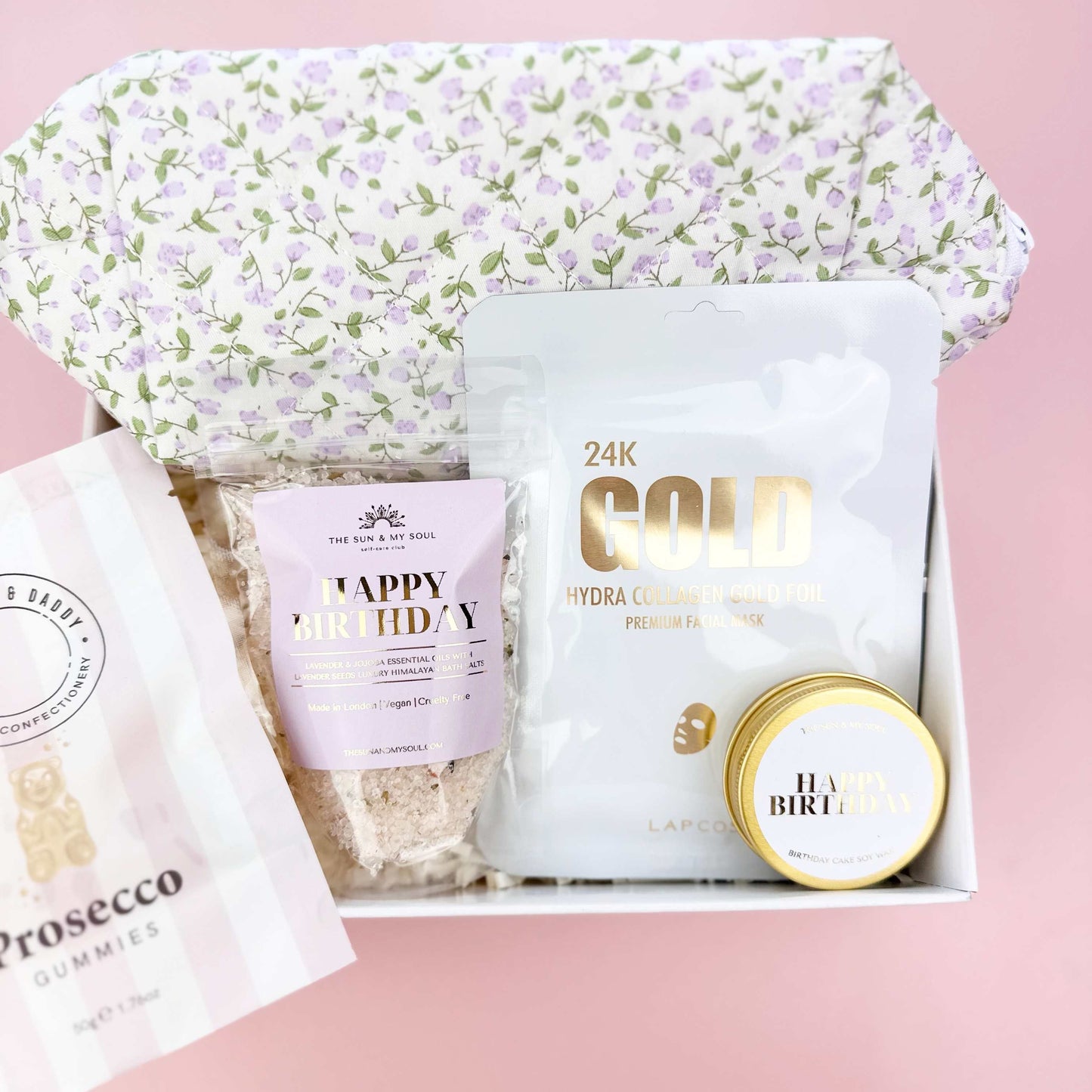 Gift set with a floral-patterned bag, gold container, and other items on a pink background
