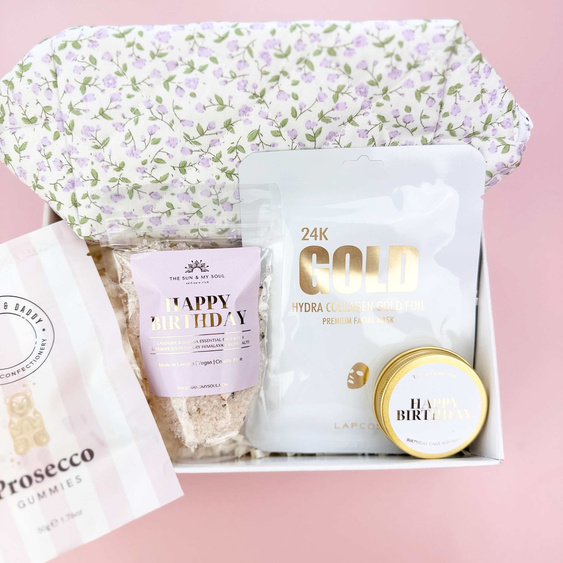 Gift set with a floral-patterned bag, gold container, and other items on a pink background