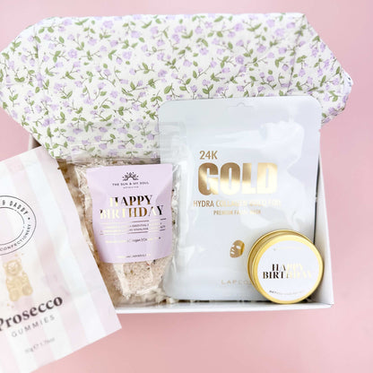 Gift set with a floral-patterned bag, gold container, and other items on a pink background