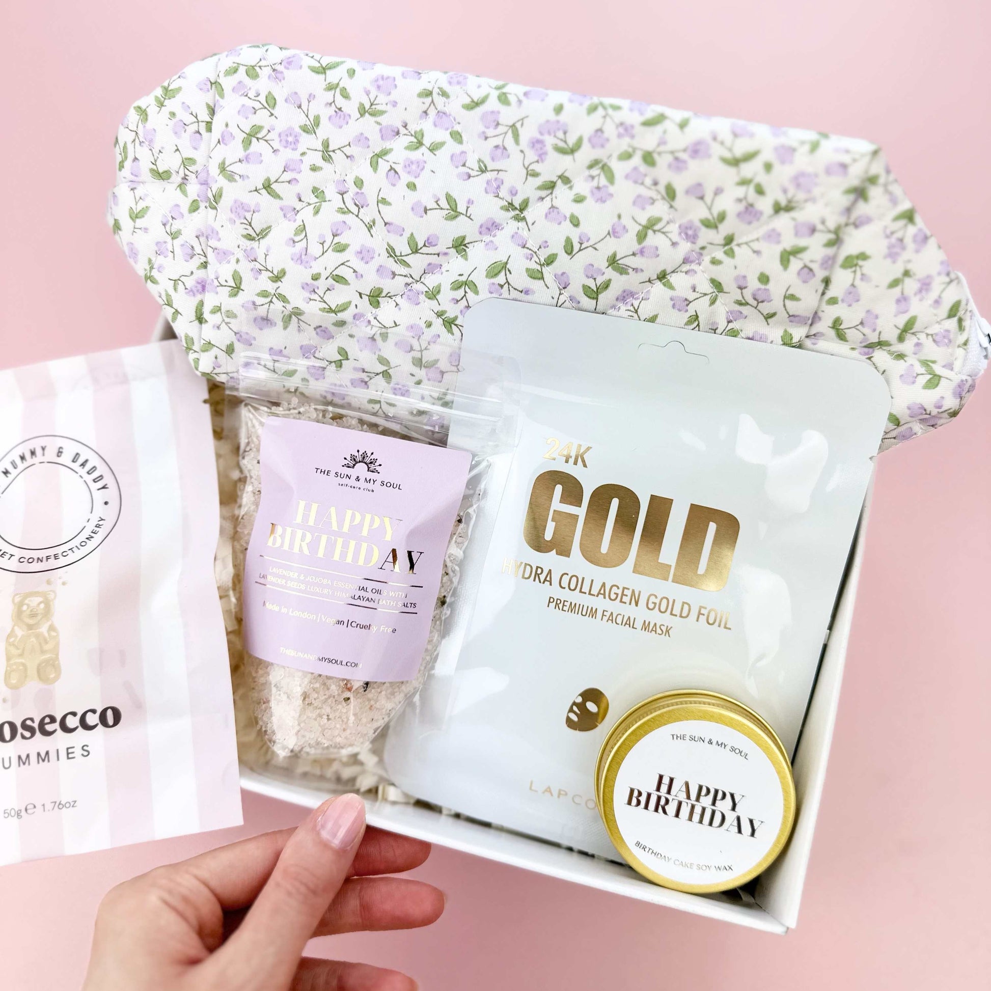 Gift box with skincare products including a gold foil pack and 'Happy Birthday' mints on a pink background.