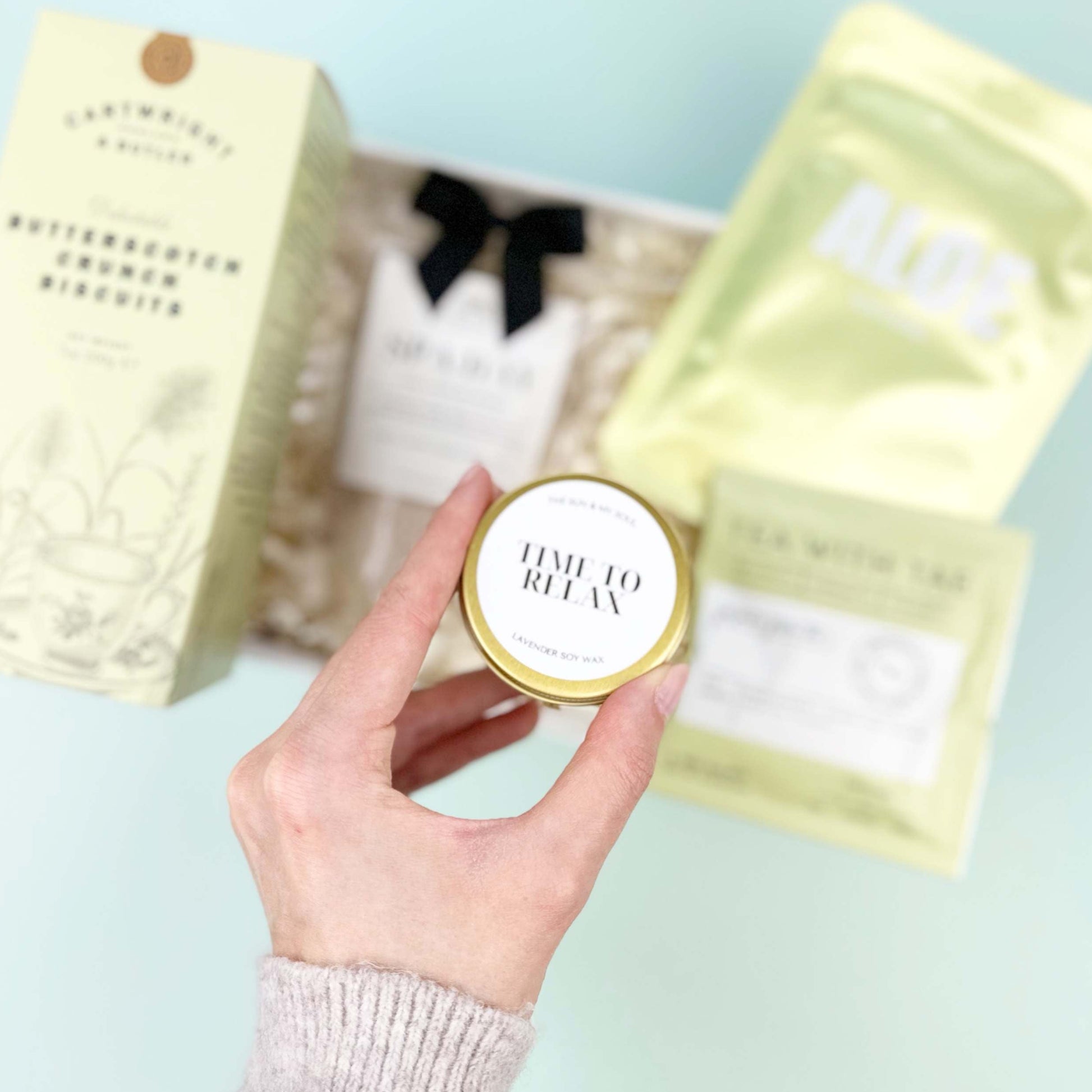 Hand holding a small round container with a lid, surrounded by larger packaging on a light green surface.