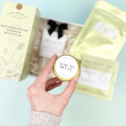 Hand holding a small round container with a lid, surrounded by larger packaging on a light green surface.