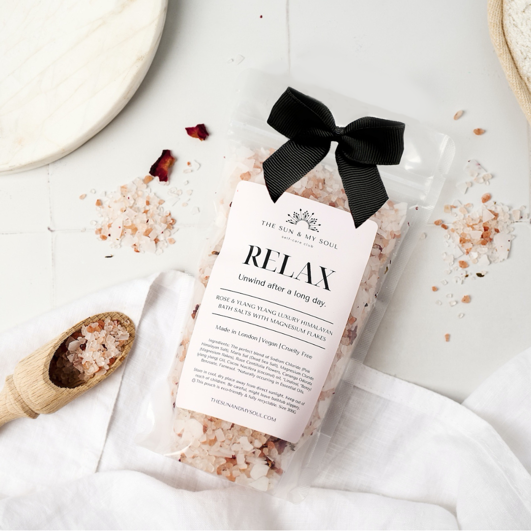 Bath salt package with a black bow on a white surface with bath salts and a wooden spoon.