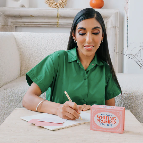 Rupi Kaur writing in a notebook with a pink 'Writing Prompts Self-Love' book on a table.