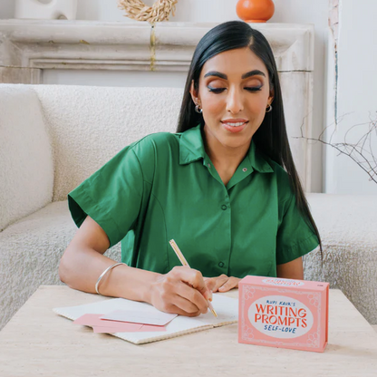 Rupi Kaur writing in a notebook with a pink 'Writing Prompts Self-Love' book on a table.