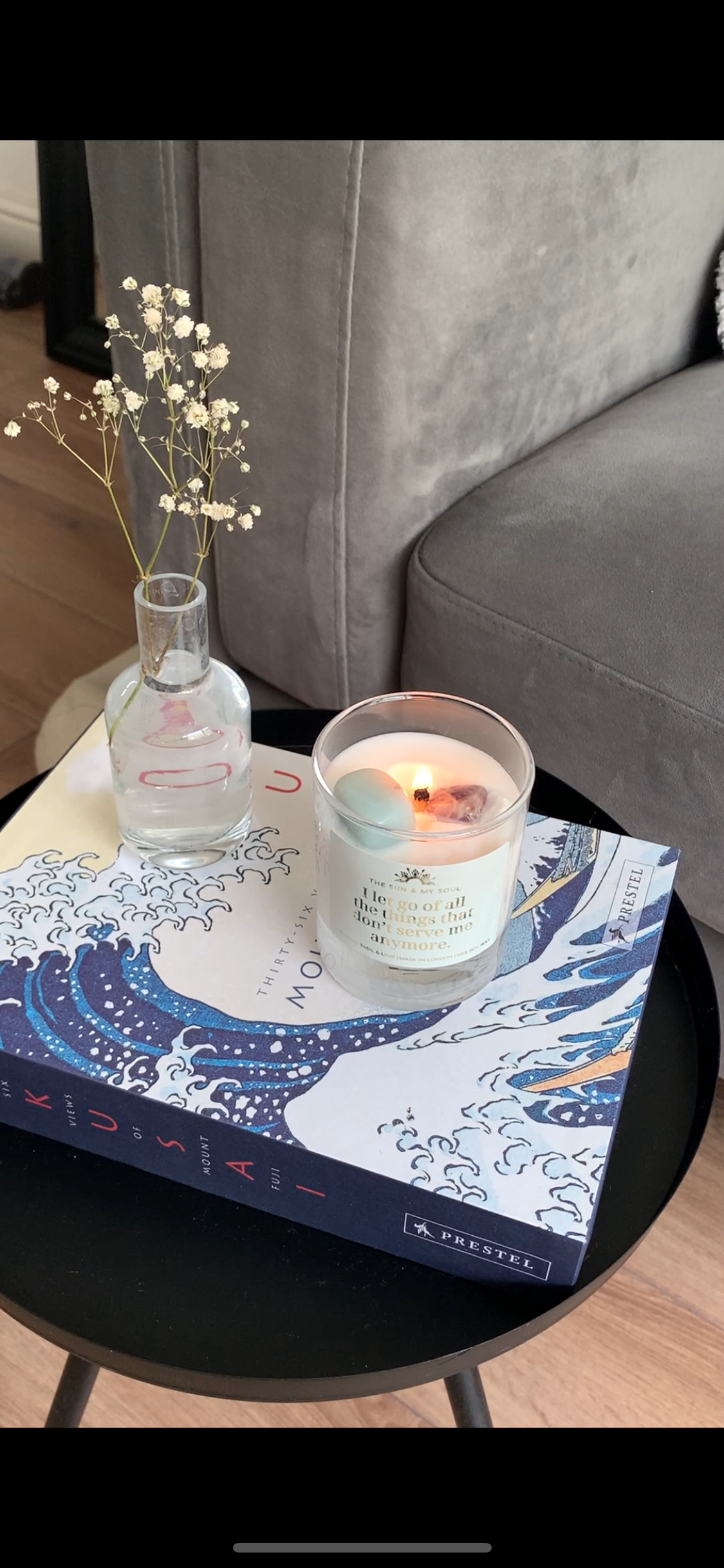 Candle on a decorative box with a vase on a small table next to a gray sofa.